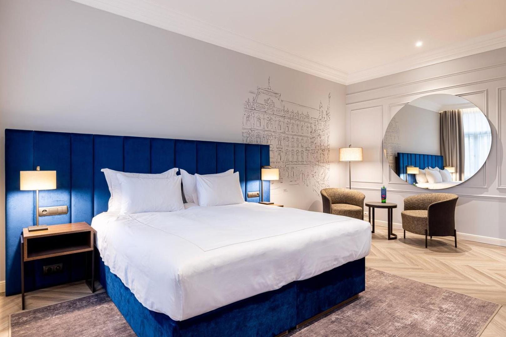 Grandhotel Pupp KLV Exterior foto The photo shows a modern hotel room featuring a large double bed with white linens and a blue upholstered headboard. The walls are light gray, and there is a decorative mural in the background. Two bedside lamps are on either side of the bed. In the 